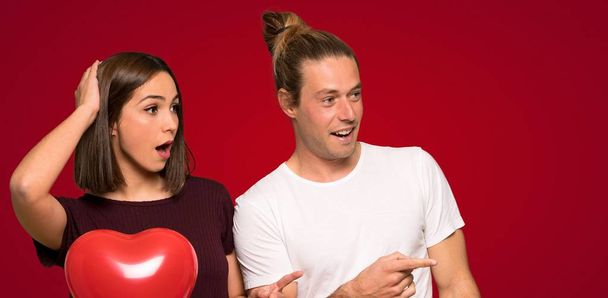 Couple in valentine day pointing finger to the side with a surprised face over red background - Foto, Imagem