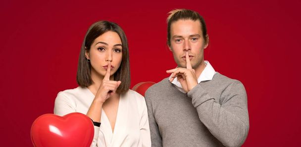 Пара в день Святого Валентина показывает знак молчания жест положить палец в рот на красном фоне
 - Фото, изображение