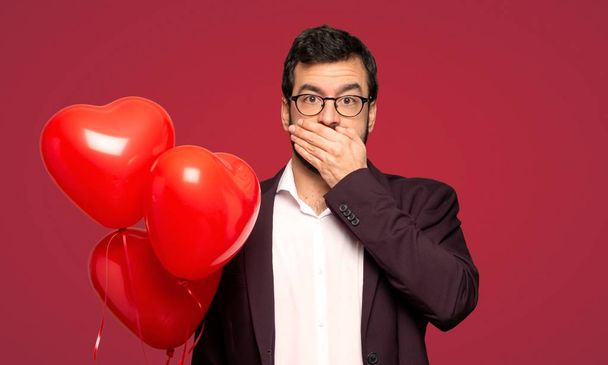 Man in valentine dag bedekking mond met handen zeg iets ongepast op rode achtergrond - Foto, afbeelding