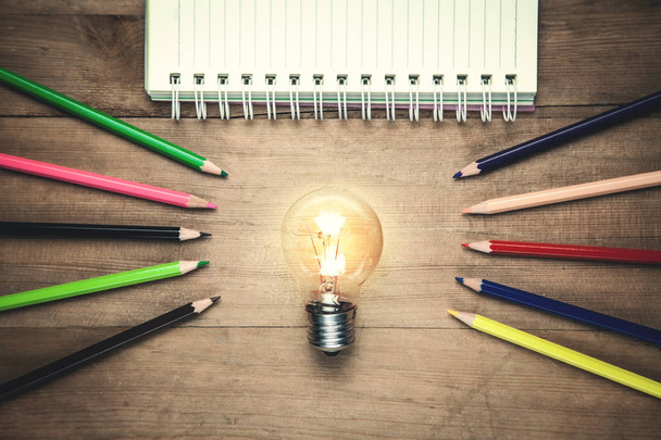 Light bulb with notepad and pencils on wooden background. - Photo, Image