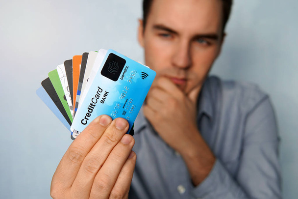 Молодой человек держит остановку кредитных и дебетовых карт в задумчивой позе. Парень выбирает карту для оплаты. бизнесмен держит в руках большую пачку кредитных карт и смотрит на них
 - Фото, изображение