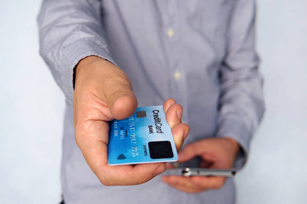 Cropped short of young man reaching out hand with blue credit card with fingerprint sensor and holding mobile in other hand. Businessman giving payment card with biometric scanner at blue background. - Photo, Image