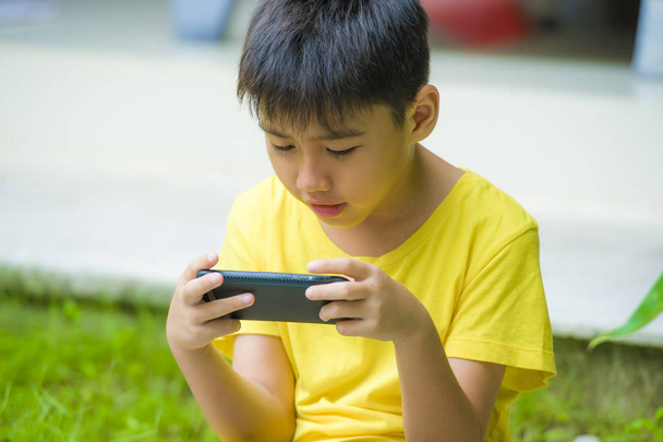 isolated lifestyle portrait of 7 or 8 years old Asian child focused and concentrated playing with mobile phone outdoors at home garden in kid suffering gaming addiction concept - Foto, Imagem