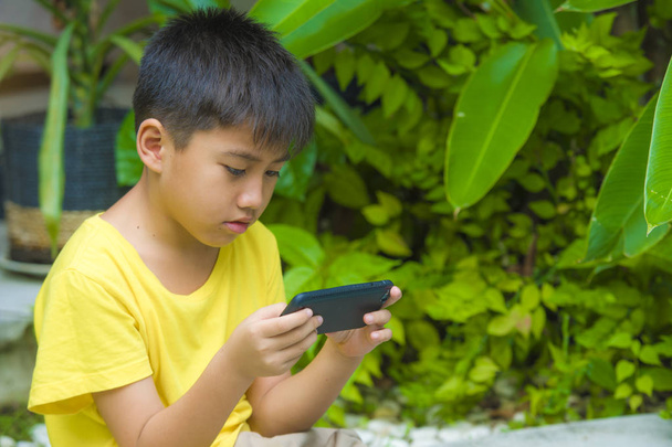 isolated lifestyle portrait of 7 or 8 years old Asian child focused and concentrated playing with mobile phone outdoors at home garden in kid suffering gaming addiction concept - Foto, Bild
