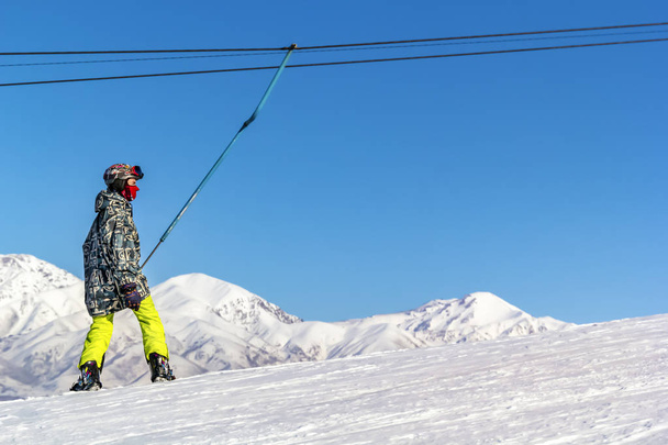 Snowboarder en équipement monte sur une montée d'ascenseur pour skier contre un ciel bleu et des sommets de montagne
. - Photo, image