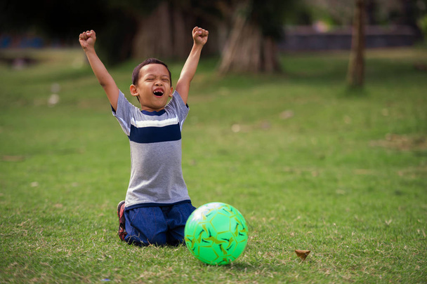 lifestyle portrait at grass city park of 5 years old Asian kid playing football happy and excited raising arms celebrating scoring goal in child sport practice education and young soccer fan concept - Photo, image