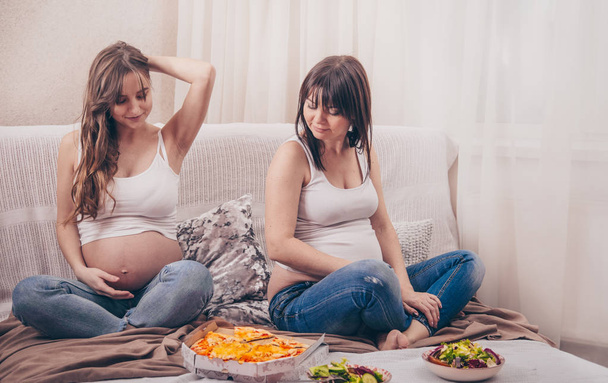 Concetto di maternità, due donne incinte che mangiano pizza e insalata a casa sul divano, amicizia e alimentazione sana
 - Foto, immagini