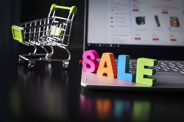 Many-colored wooden word SALE set on laptop bottom at the background of opened web page on the screen and small shopping trolley. Concept of shopping online. Good buy in internet. Sale in E-commerce. - Photo, Image