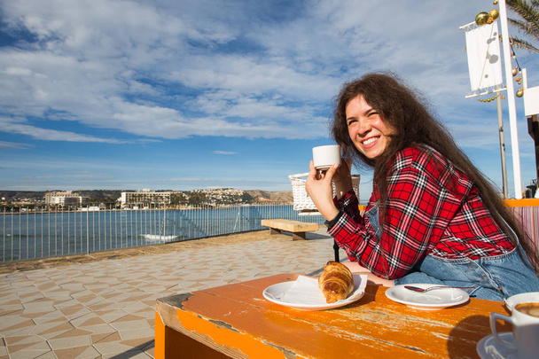 Jeune femme petit déjeuner avec croissant et café au café dans la rue en face de la mer
 - Photo, image