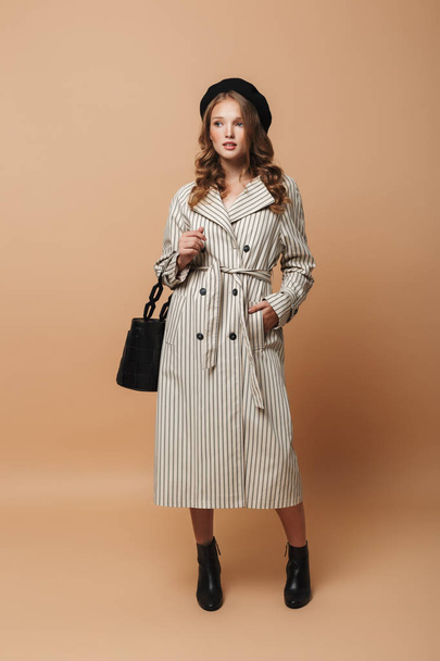 Young attractive woman with wavy hair in striped coat and black hat holding bag in hand dreamily looking aside over beige background - Foto, Bild