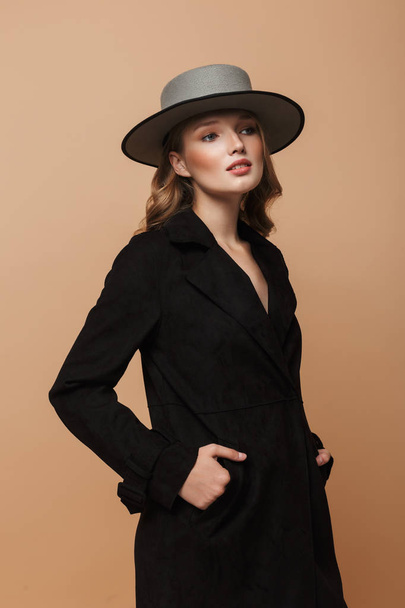 Jovem mulher linda com cabelo ondulado em casaco preto e chapéu cinza sonhadoramente olhando para o fundo bege
 - Foto, Imagem