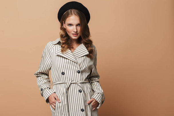Young gorgeous woman with wavy hair in striped coat and black hat thoughtfully looking in camera over beige background - Photo, Image