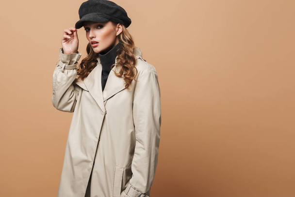Young beautiful woman with wavy hair in classic trench coat and cap thoughtfully looking in camera over beige background - 写真・画像