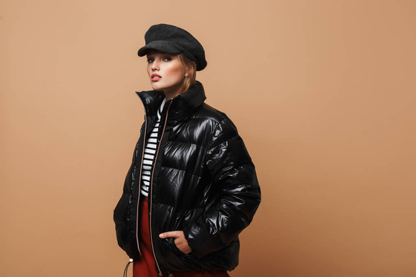 Young beautiful serious woman with wavy hair in cap and black down jacket thoughtfully looking in camera over beige background - Фото, зображення