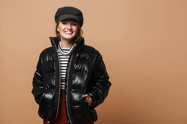 Young beautiful smiling woman with wavy hair in cap and black down jacket joyfully looking aside over beige background - Foto, afbeelding