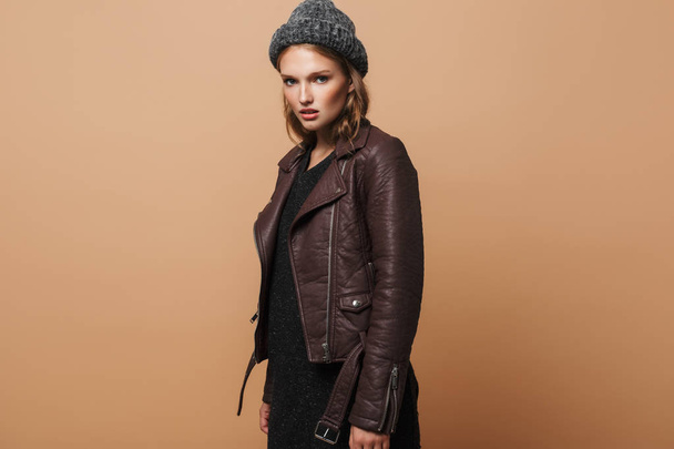 Young attractive serious woman in cap,leather jacket and black dress thoughtfully looking in camera over beige background - 写真・画像
