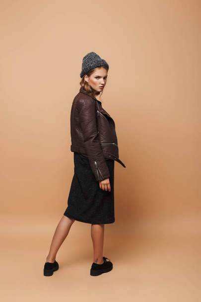 Young pensive woman in hat,leather jacket and black dress angrily looking in camera from back over beige background - Photo, image