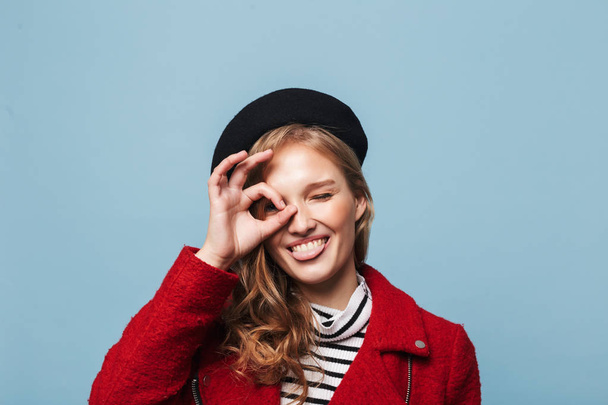 Close-up van de mooie lachende meisje met golvend haar in zwarte baret en een rood jasje speels tong in de camera kijkt uit over de blauwe achtergrond weergegeven: - Foto, afbeelding