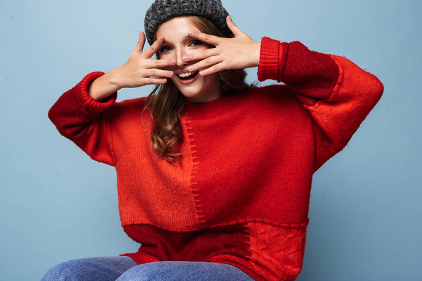 junge schöne lächelnde Frau mit welligem Haar in Hut und rotem Pullover, die glücklich in die Kamera schaut, das Gesicht mit den Händen vor blauem Hintergrund verdeckt - Foto, Bild