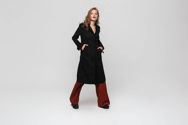 Young gorgeous woman with wavy hair in black coat and red wide trousers dreamily looking in camera over gray background - Fotoğraf, Görsel