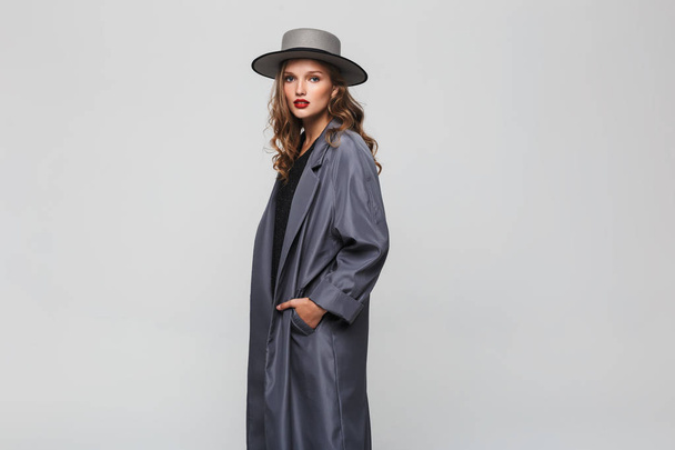 Young gorgeous woman with wavy hair and red lips in hat and cloak holding hands in pockets dreamily looking in camera over gray background - Photo, Image