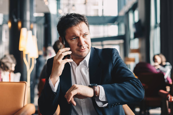 Portrait of young handsome lawyer checking the time on his hand watch and having a phone conversation, while sitting in a modern restaurant during a business break. Time is money concept. - Photo, Image