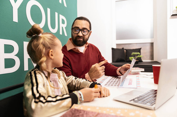 Dedicated teacher. Dark-haired bearded teacher in a brown sweater and eyeglasses looking involved while showing pictures on laptop to his pupil - Foto, imagen
