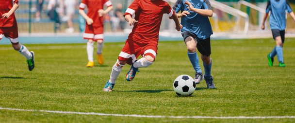 Labdarúgás futball játékosok fut a labdával. Labdarúgók rugdossa a labdarúgó-mérkőzés. Fiatal focisták futás után a labdát. Gyerekek Labdarúgás piros és kék egyenruhát. Labdarúgó-stadion, a háttérben  - Fotó, kép
