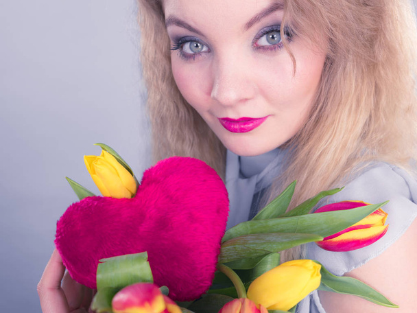 International womens or valentines day. Attractive grateful woman blonde hair holding tulips bunch and red heart sign. On grey - 写真・画像
