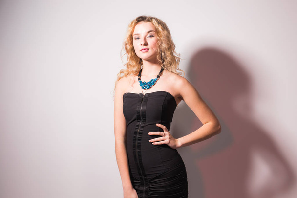 Fashion and model concept - Beautiful young sexy blond woman in a black dress posing against a white wall - Photo, Image