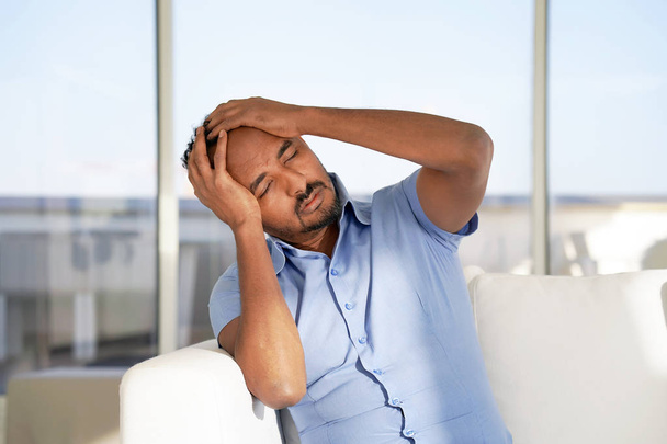 Mann leidet unter Kopfschmerzen Migräne Schmerzen zu Hause auf dem Sofa. Gesundheitsprobleme, Stress und Depressionen. Afrikanischer Rüde hält Kopf mit Hand. Gesundheitskonzept. - Foto, Bild