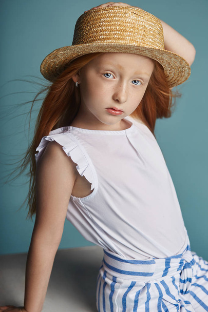 Norwegian girl with bright red hair in a straw hat. Beautiful redhead baby girl with long hair and beautiful big blue eyes. Red-haired girl child in summer clothes posing on a blue background - Foto, Imagen