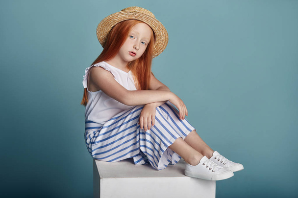 Norwegian girl with bright red hair in a straw hat. Beautiful redhead baby girl with long hair and beautiful big blue eyes. Red-haired girl child in summer clothes posing on a blue background - Foto, afbeelding