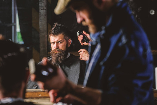 Barber with hair clipper works on hairstyle for man with beard, barbershop background. Haircut concept. Hipster client getting haircut. Barber styling hair of brutal bearded client with clipper - Foto, afbeelding