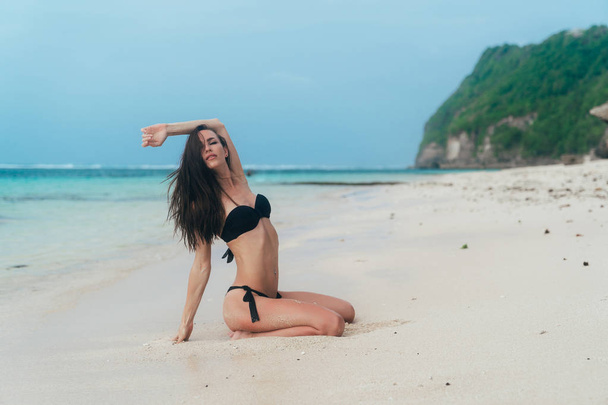 Sexy brunette girl in black swimwear sits and sunbathes on ocean beach - Foto, Bild