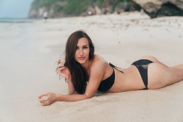 Beautiful tanned brunette girl in swimsuit lying on sandy beach. Model with sexy booty posing near ocean - Fotoğraf, Görsel