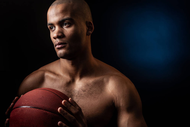 Studio portrait of Black basketball player holds a ball over grey background. - Фото, изображение