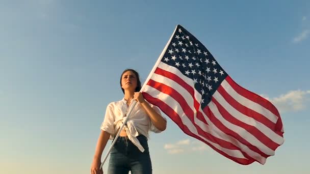 Chica seria adolescente de pie con bandera americana contra el cielo azul. Movimiento lento
 - Metraje, vídeo