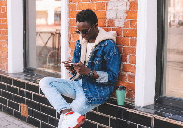 Fashion cool urban african man with smartphone sitting on city street, brick wall background - Фото, зображення