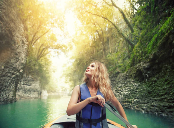 azure mountain river glistens in sun, charming happy smiling girl with lush blond hair sits in boat on lake in Georgia, nature Martvili Canyon, rafting as active leisure for tourists, bright sunshine - Foto, Imagem
