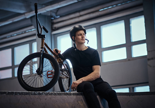 Bmx rider relaxing after practicing tricks with his bike in a skatepark indoors - Photo, Image