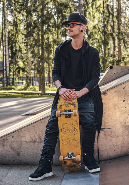 Elegante skateboarder en una sudadera con capucha negra y gorra se sienta en la rampa en el skatepark en la hora de verano
 - Foto, Imagen