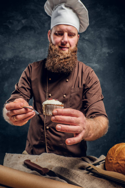Het dragen van een uniform houden van een heerlijke muffin in een donkere studio Baker. - Foto, afbeelding