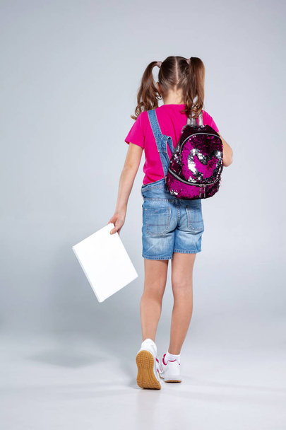 Schoolgirl in glasses with backpack and textbook on the gray background - Photo, Image