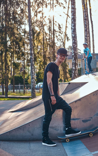 Стильный скейтбордист в черной рубашке и кепке, стоящий на доске в скейтпарке летом
 - Фото, изображение