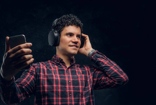 Vezeték nélküli fejhallgató fiatalember zenét hallgat, és tartja a smartphone egy stúdióban, a háttérben a sötét fal - Fotó, kép