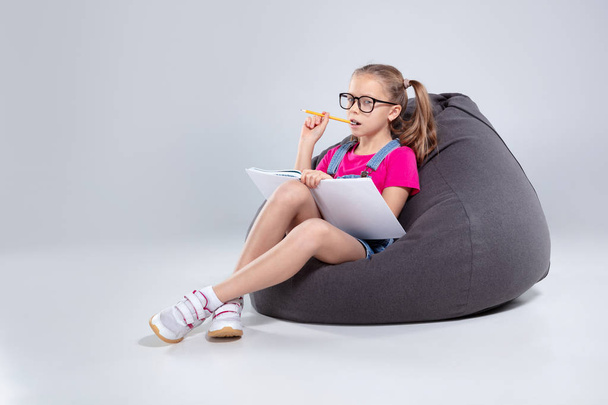 Young girl in glasses doing homework on a gray bean bag - Foto, immagini