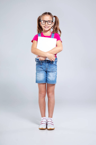 Schoolgirl in glasses with backpack and textbook on the gray background - Фото, изображение