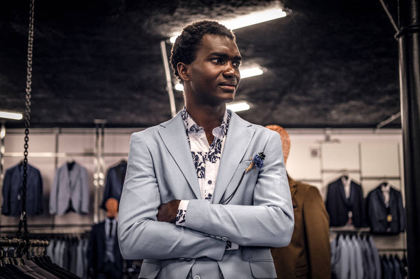 Uomo afro-americano vestito di blu elegante vestito in posa con le braccia incrociate in un classico negozio di abbigliamento maschile
. - Foto, immagini