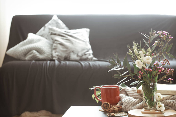 Acogedora sala de estar interior de la casa con un sofá negro y un jarrón con flores y artículos de decoración en una pequeña mesa
 - Foto, Imagen